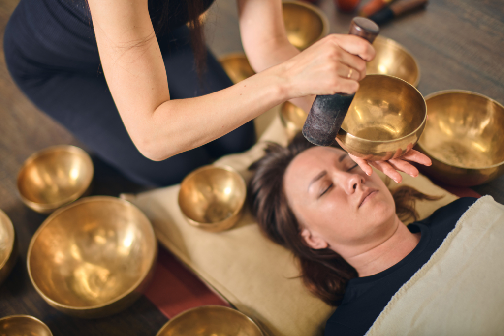 Young woman lies on floor surrounded by golden singing bowls. Reiki practitioner uses mallet to create soothing vibrations. Calm, meditative atmosphere prevails