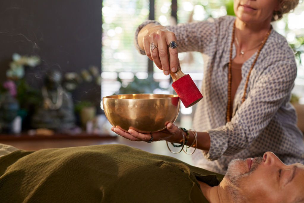 Close up hand of mature woman using singing bowl for sound healing on relaxed middle aged man at wellness center. Vibrational therapy session in peaceful space. Mature woman therapist performing sound therapy on senior man lying on back.
