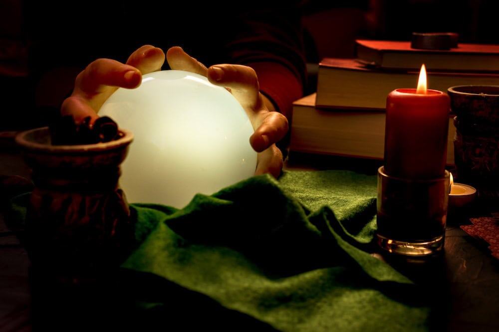 A pair of hands rests on a large, glowing white crystal ball in a dimly lit room. To the right, a thick red candle burns next to a stack of books, with the soft light reflecting off a green velvet cloth on the table.