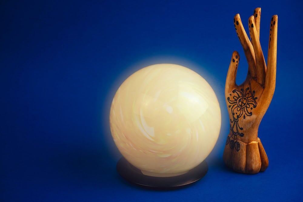 A luminous, glowing white crystal ball sits on a dark circular stand against a vibrant blue background. To the right stands a wooden decorative hand adorned with intricate black floral henna-style patterns.