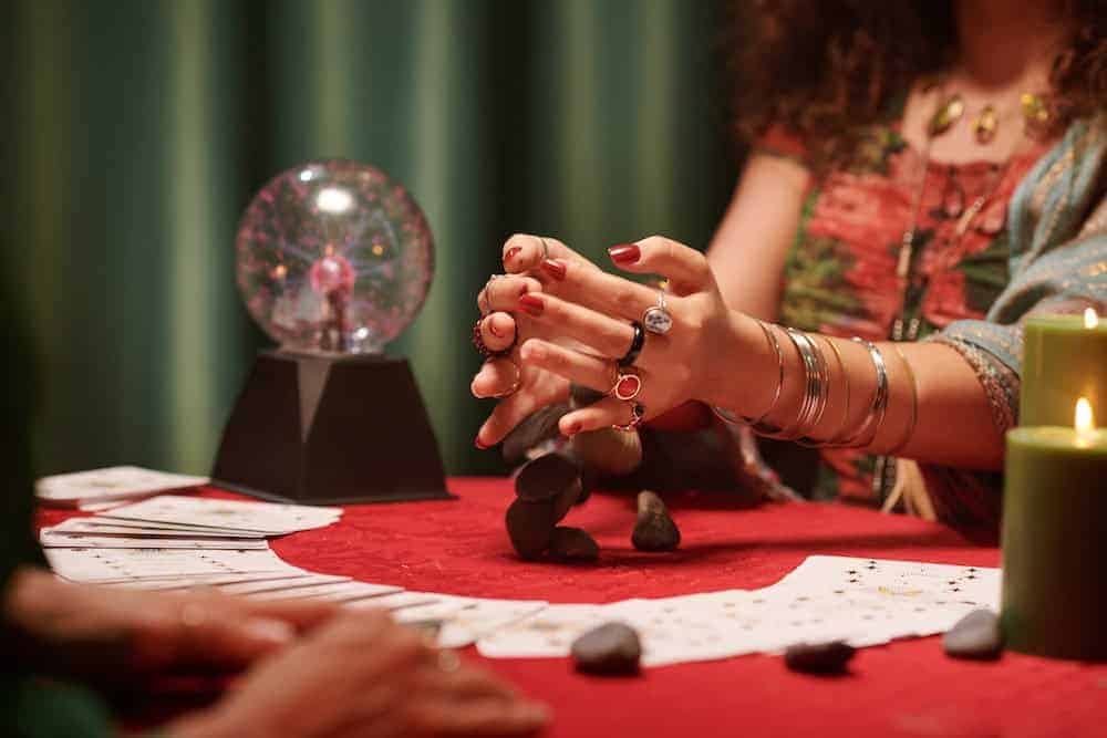 A woman with many rings and bracelets on her hands is shown mid-action casting small stones or pebbles onto a red tablecloth. The scene includes fanned-out tarot cards, a plasma ball on a black stand, and lit green candles in the background.