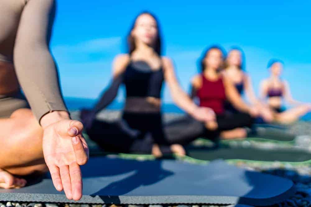 A close-up of a person's hand in a mudra during a group meditation session on a rocky beach, with other participants blurred in the background.