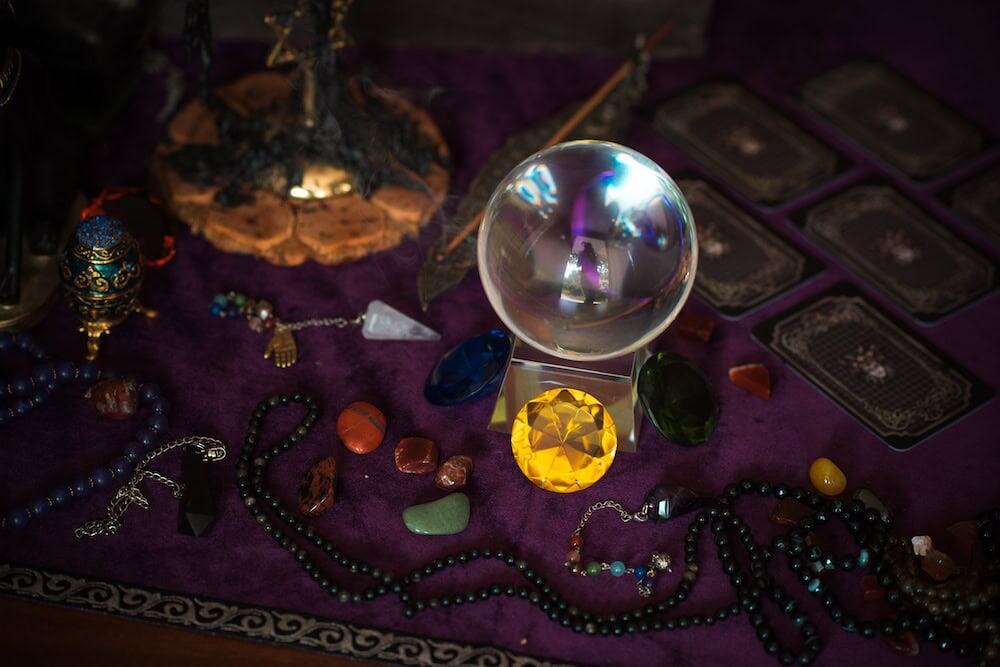 A clear crystal ball sits centrally on a stand over a dark purple velvet cloth, surrounded by various face-down tarot cards, colorful tumbled gemstones, a large faceted yellow gem, beaded necklaces, and a pendulum.