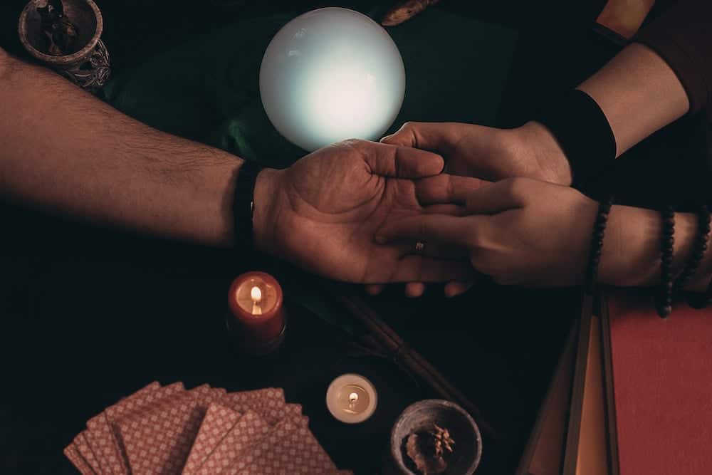 A top-down view of a palmist tracing lines on a person's hand during a reading. The scene is illuminated by a glowing white orb in the center, a lit red candle, and a small tealight, with a deck of patterned cards visible in the corner.