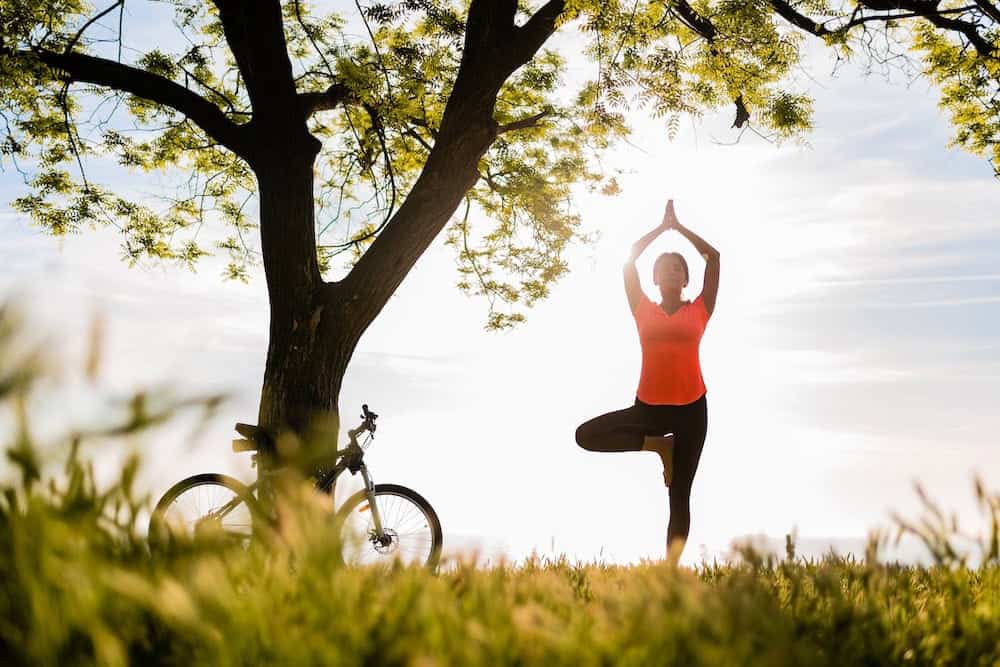 A woman in a bright orange top performs the Tree Pose (Vrksasana) under a large, leafy tree next to her bicycle during a sunny day.