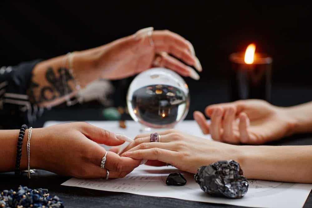 A close-up photograph of a palm reading session where a person with tattooed arms and long white fingernails holds the hand of another person. The table setting includes a clear crystal ball, a lit black candle, and various dark gemstones.