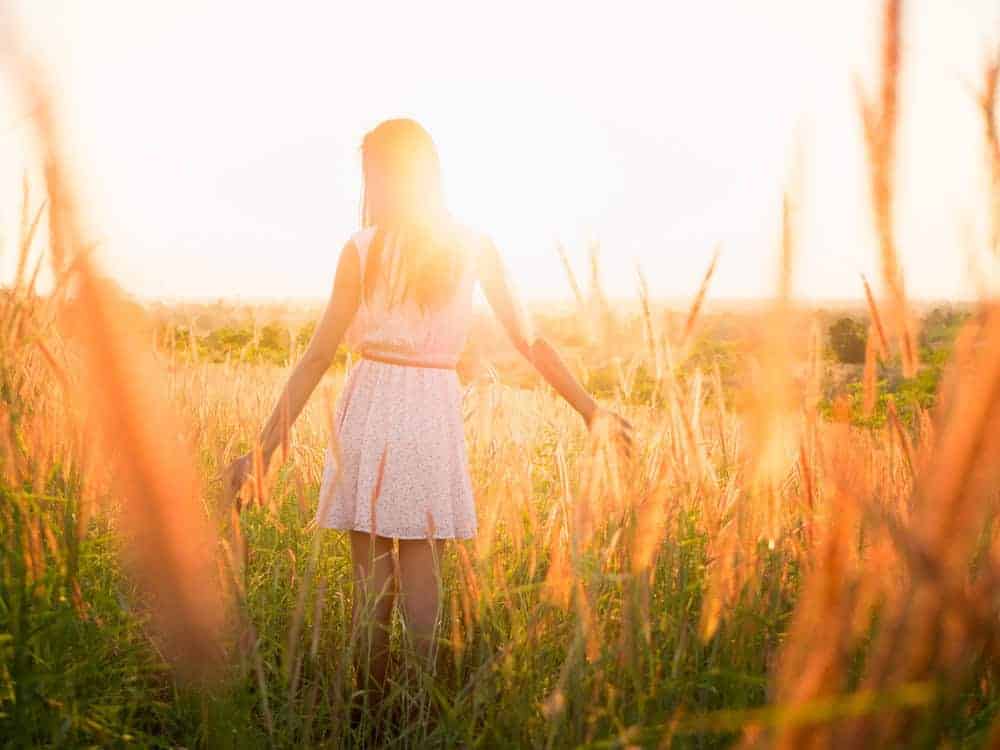 A woman in a light summer dress walks away from the camera through a sun-drenched field of tall grass during a bright sunset.